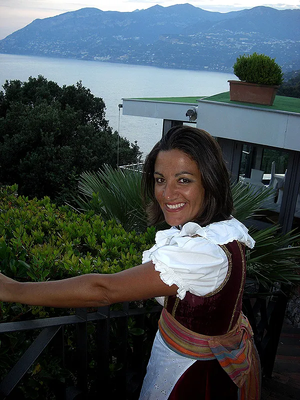 A dancer in a burgundy bodice and striped sash smiling on a terrace above the bay