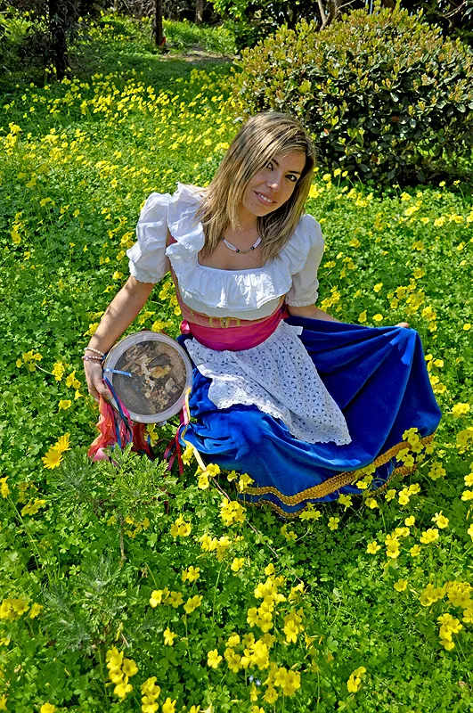A dancer in costume kneeling in a meadow of yellow flowers, with a blue skirt and tambourine