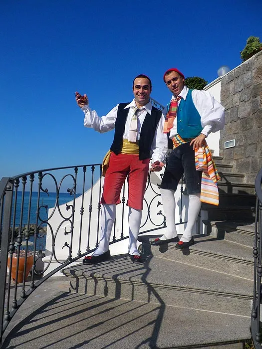 Two male dancers in costume posing on a stairway above the sea under a deep blue sky