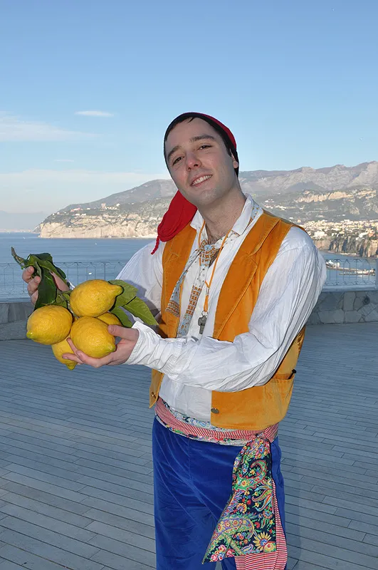 A dancer in an orange waistcoat and red cap holding out a lemon branch, the Sorrento coast behind