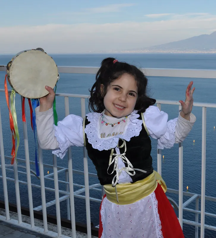 A little girl in traditional costume smiling as she raises a tambourine on a terrace above the sea
