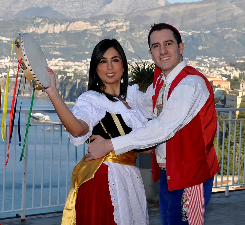 A couple in costume embracing with a ribboned tambourine, the Sorrento coastline behind them