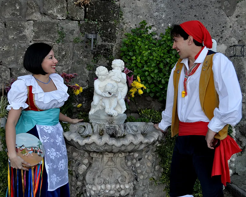 A couple in costume on either side of a fountain with stone cherubs, among flowers and old walls