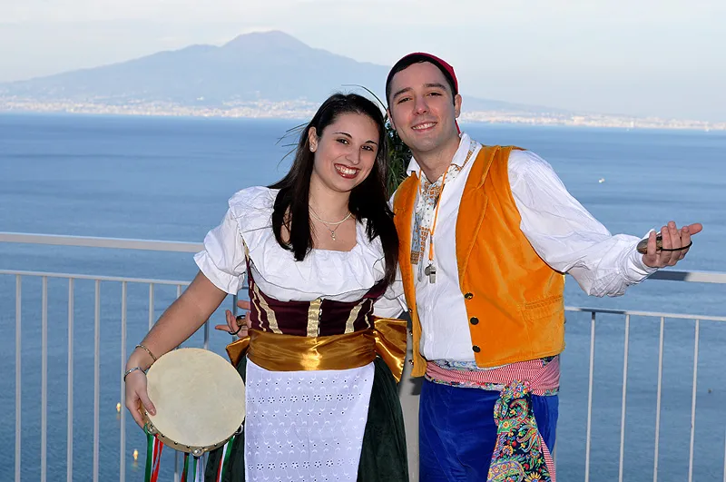 A couple in costume on a terrace, he in an orange waistcoat, she with a tambourine, Vesuvius behind