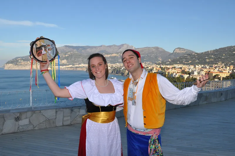 A smiling couple in costume, her tambourine raised, on a lookout with Sorrento behind