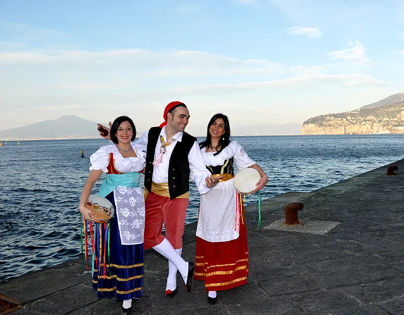 A male dancer between two female dancers in costume on a pier, the bay and Sorrento coast behind