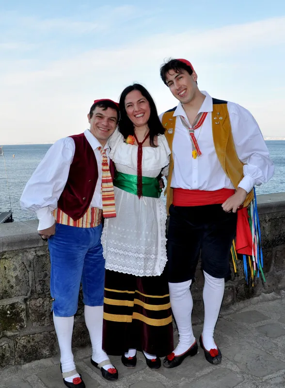 Two male dancers and one female dancer in costume arm in arm by a sea wall