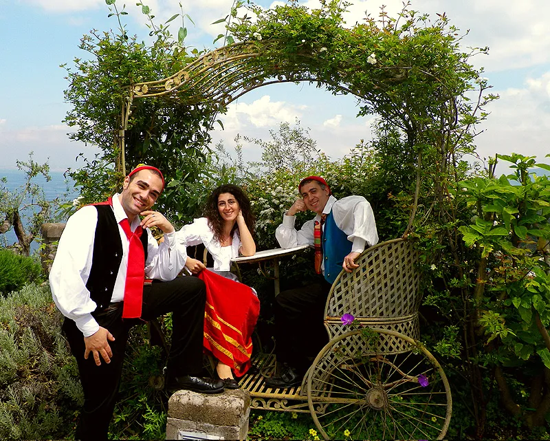 Three dancers in costume posing under a flowering arch beside a vintage carriage