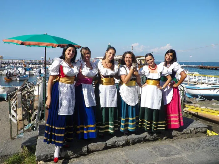 Six female dancers in costume posing at the harbour under an umbrella, boats behind them