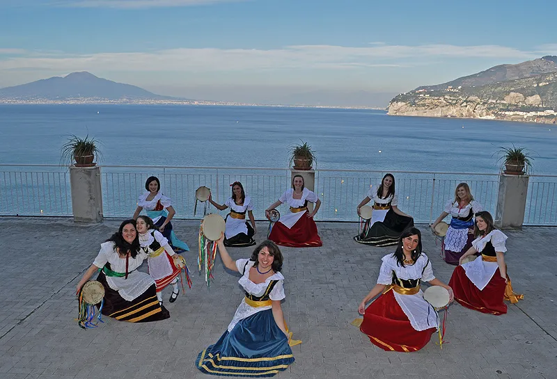 Dancers in costume seated in a fan shape on a terrace, with Vesuvius and the coast behind