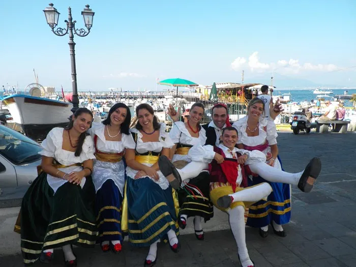 Dancers in costume sitting on a harbour wall, one playfully lying across their laps