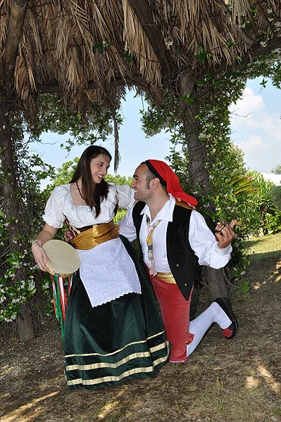 A kneeling dancer courts his partner, who holds a tambourine, under a thatched hut