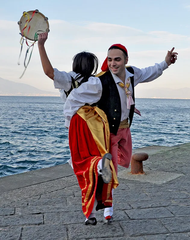 Coppia danza la tarantella sorrentina su un molo, lei con il tamburello alzato verso il cielo, il mare alle spalle
