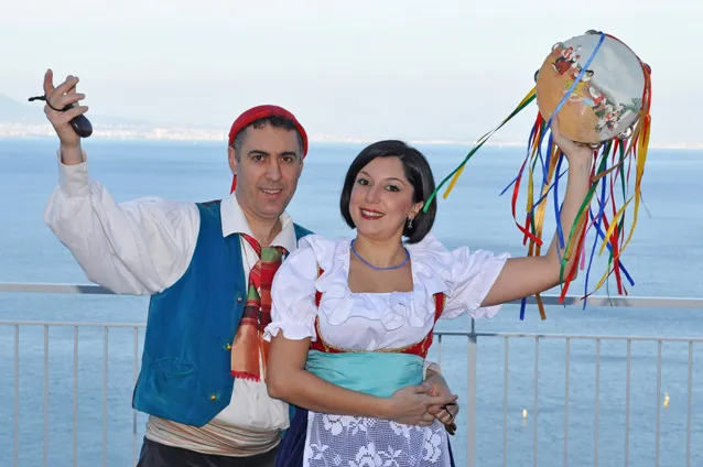 A smiling couple in costume with a ribboned tambourine and castanets in front of the sea