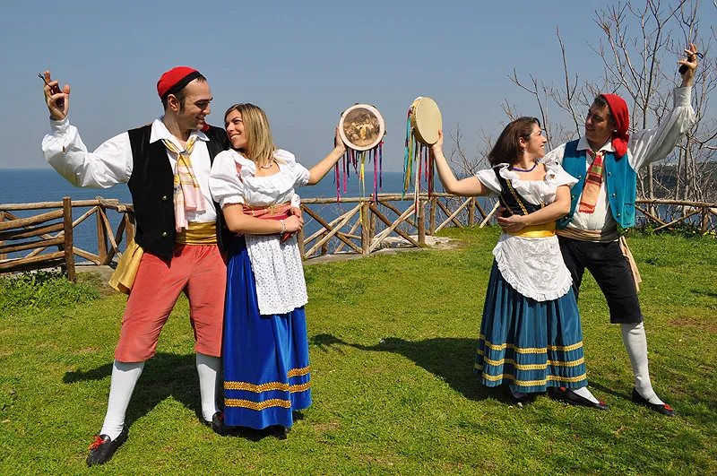 Two couples in costume dancing with tambourines on a lawn above the sea