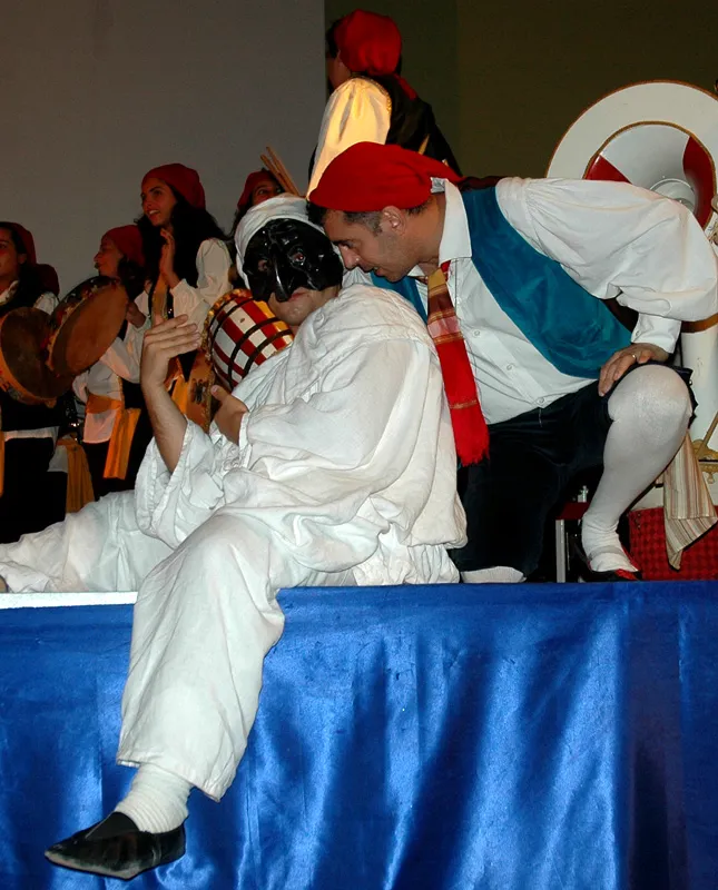 Pulcinella sitting on the edge of the stage among the dancers during a tarantella show